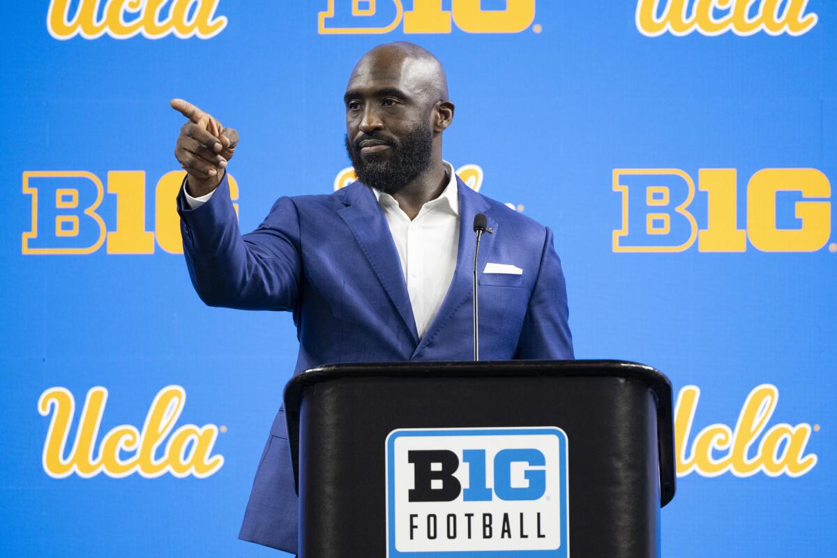 UCLA coach DeShaun Foster stands at a podium and points while talking with reporters 