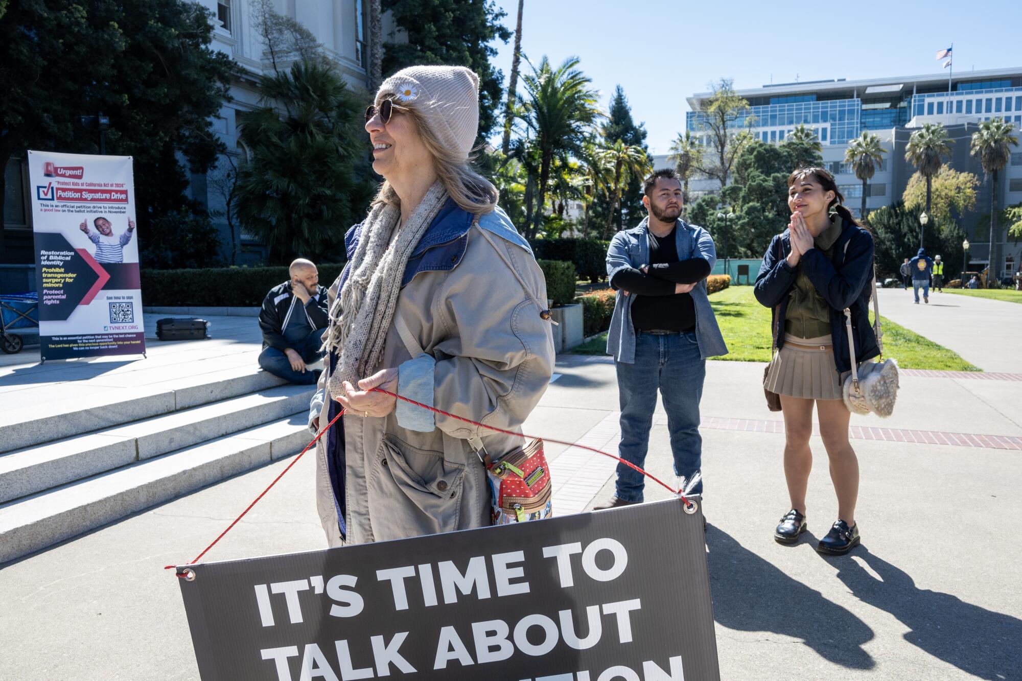 A woman holds a sign and a man and a woman stand and talk nearby. 