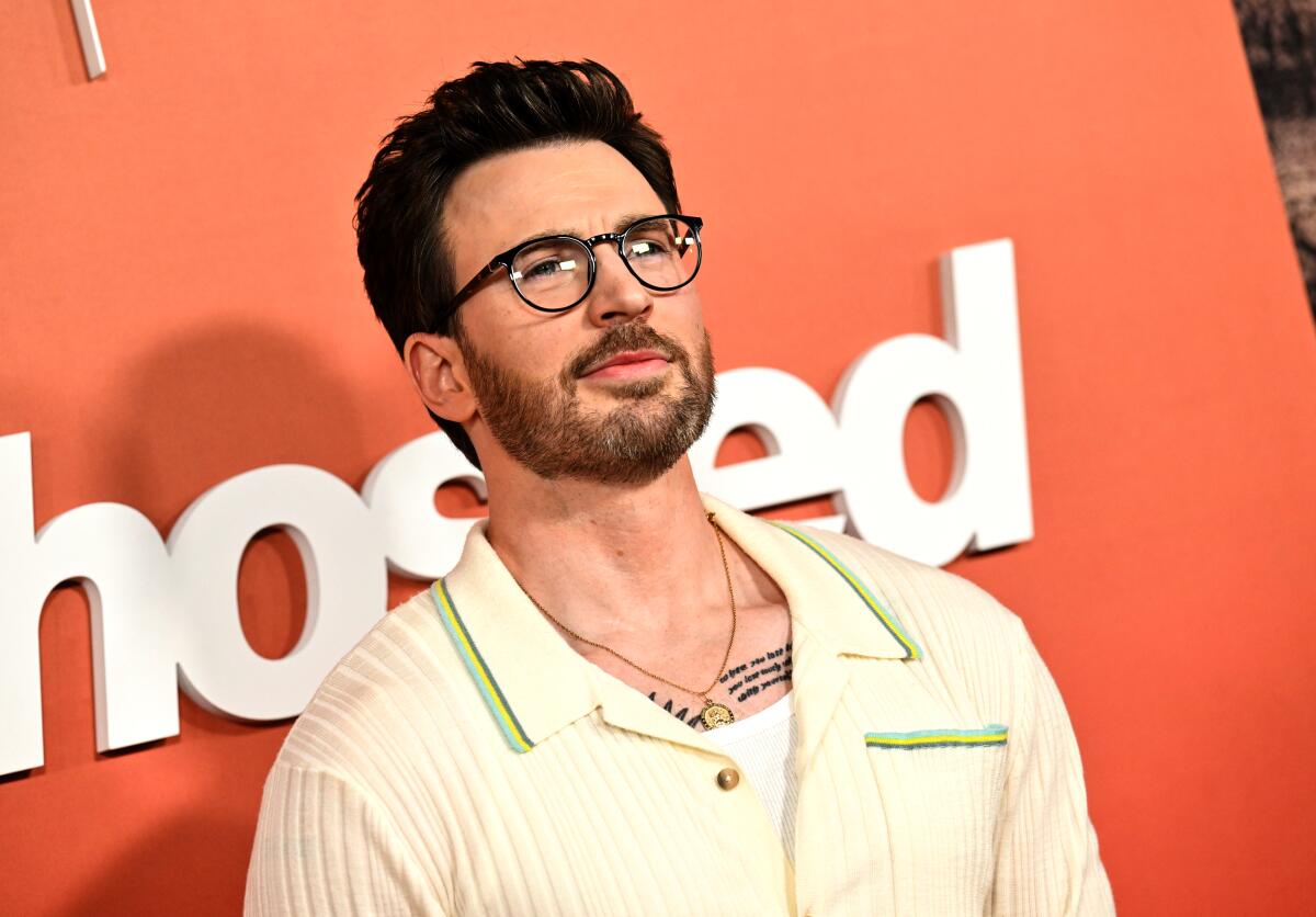 Chris Evans wears glasses in front of an orange backdrop.