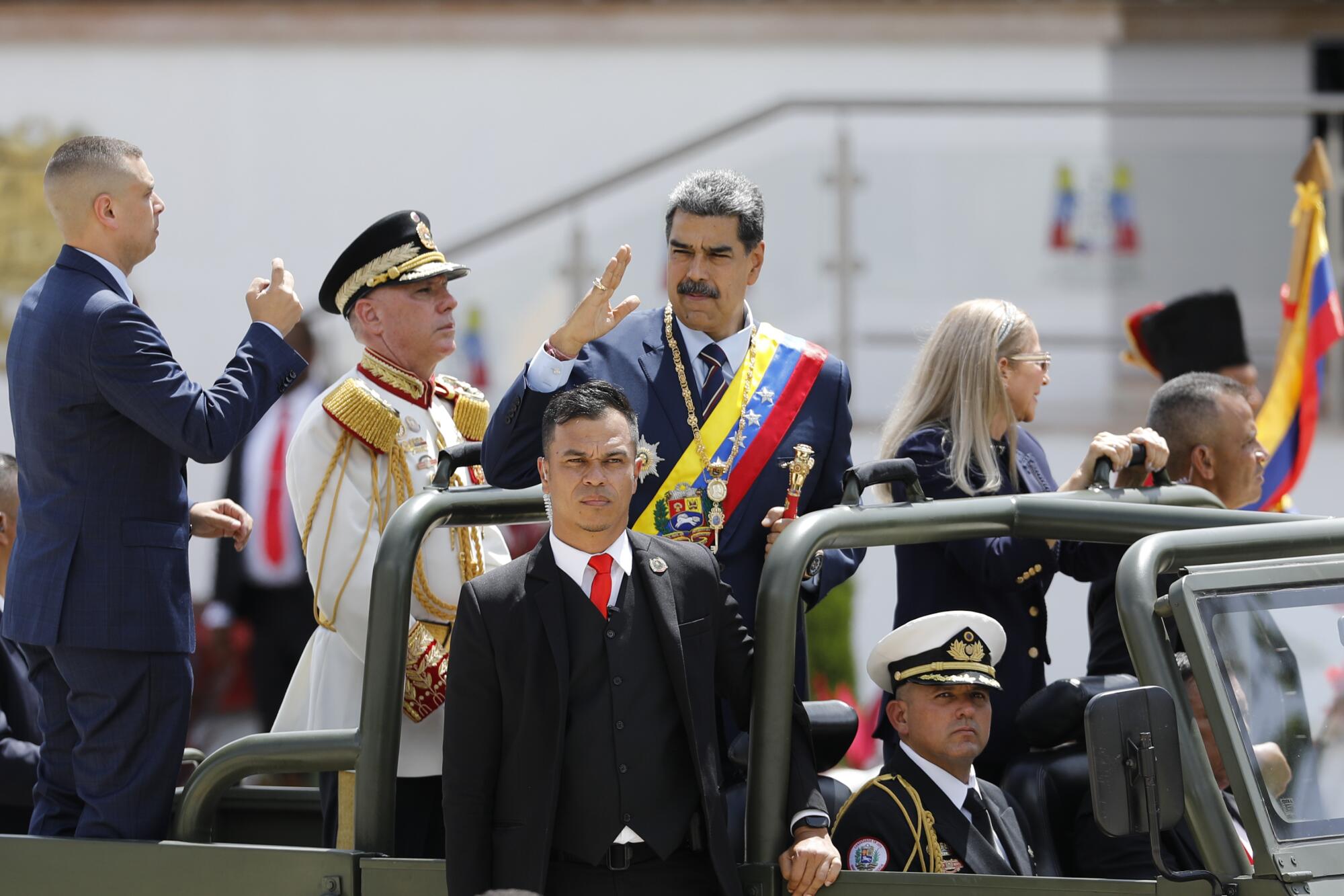 A man with dark hair and mustache, with a blue, yellow and red sash over his dark suit, salutes while standing in a vehicle 