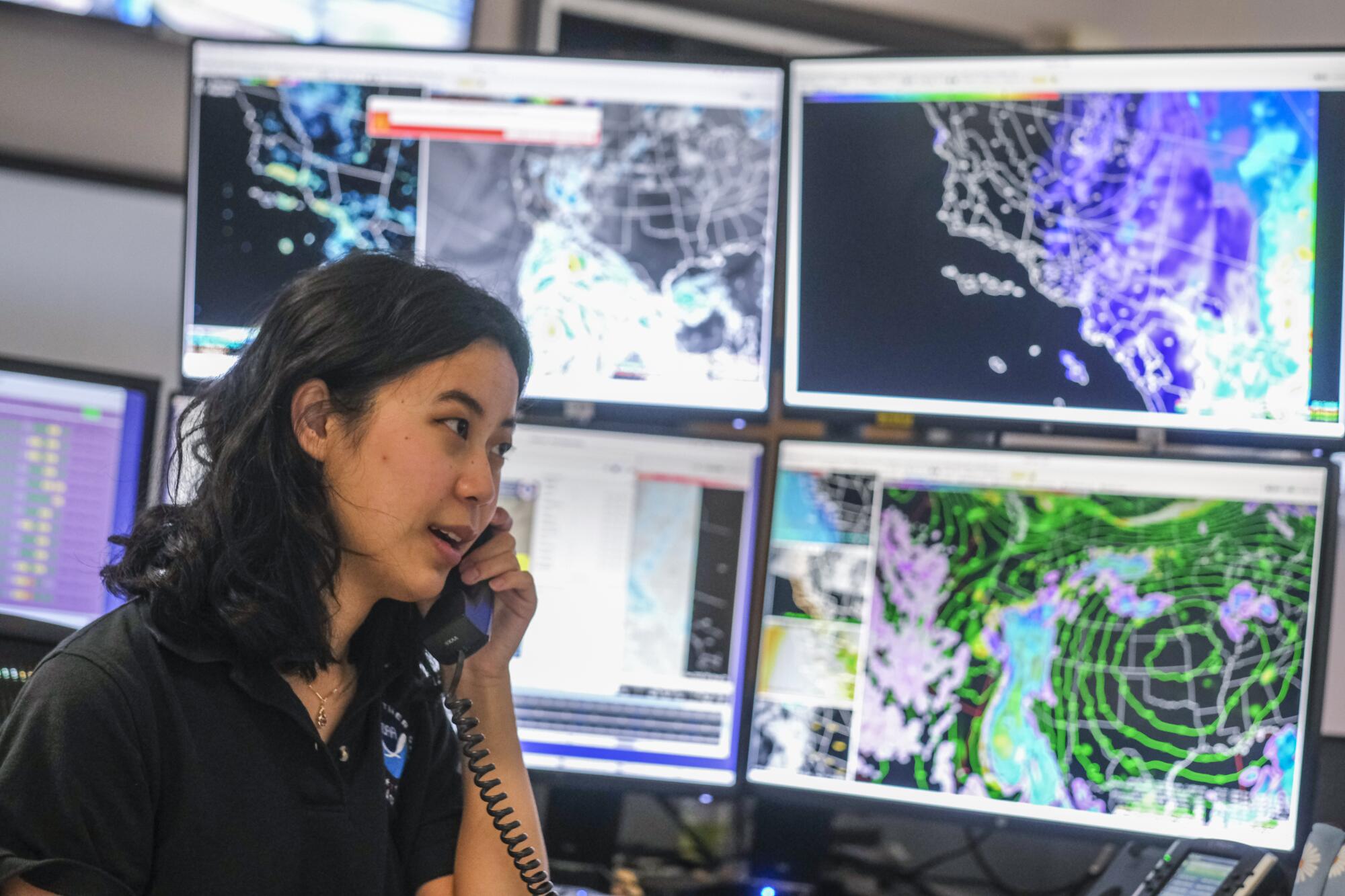 A woman talks on the phone near a bank of computer monitors displaying weather data.