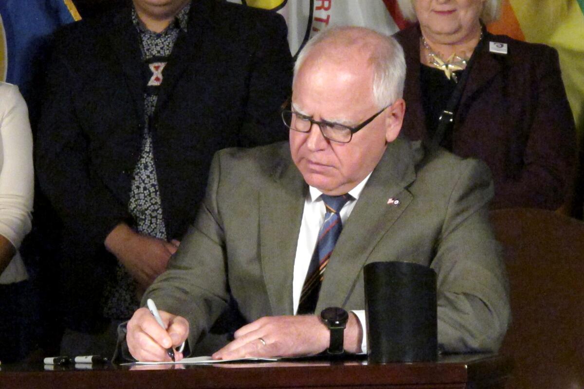 A man with gray hair, in glasses and gray jacket, signs a document as other people stand behind him