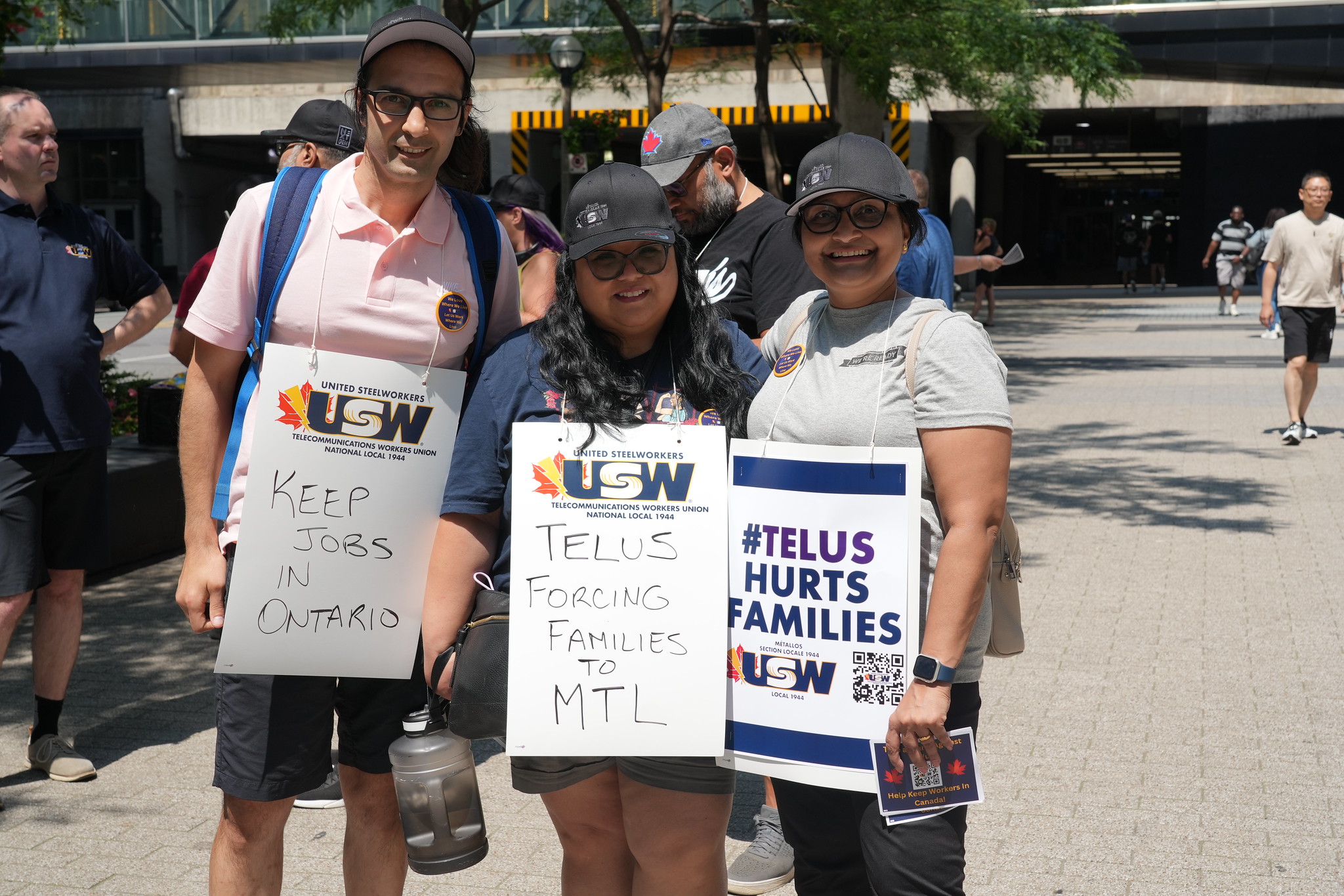 Image: three people stand outdoors on a sunny day wearing placards: