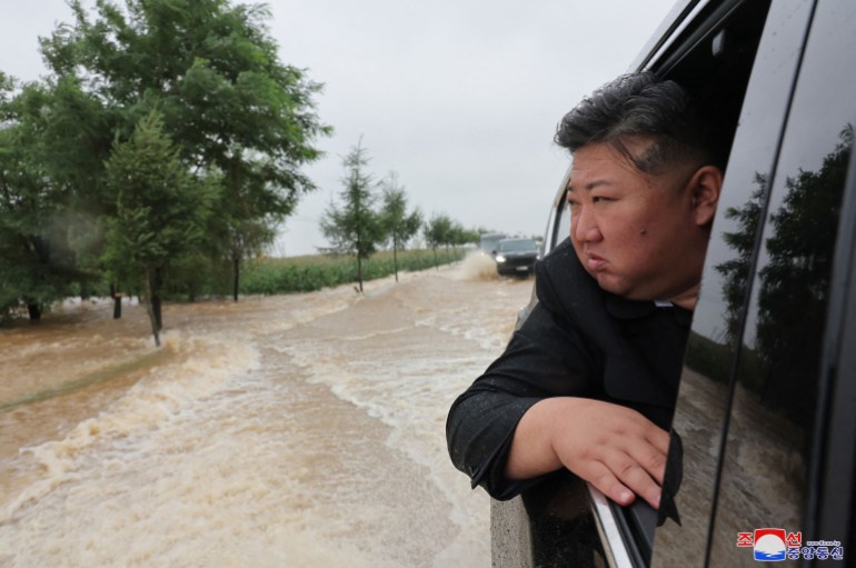 Kim Jong Un looking out of a black car. The vehicle is being driven through brown flood waters. Kim looks serious.