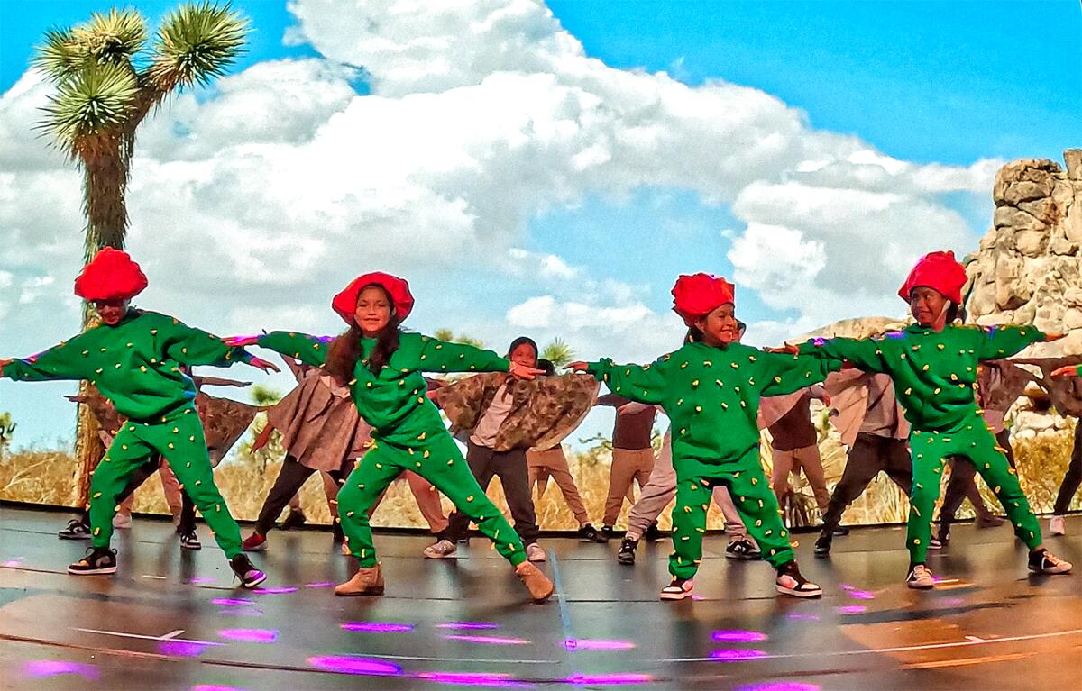 Kids dressed as cactus with a red flower hat link arms onstage.