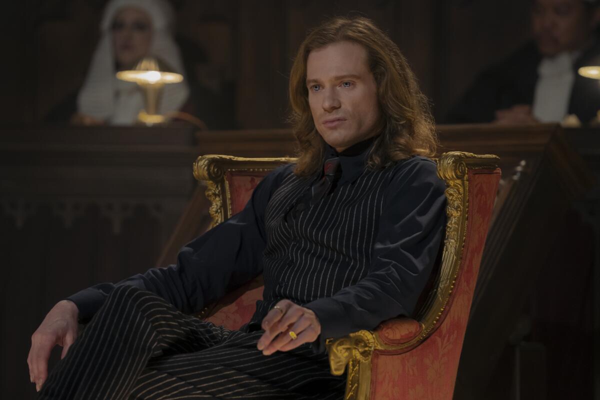 A man with long hair and dark clothing sitting on an orange velvet chair.