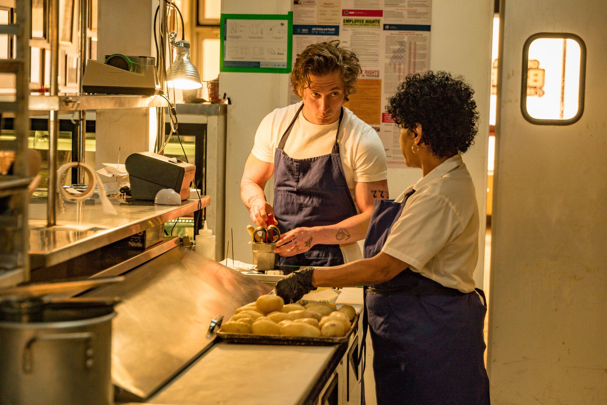 A man and a woman, both wearing a blue aprons, stand in a restaurant kitchen as they prepare food.