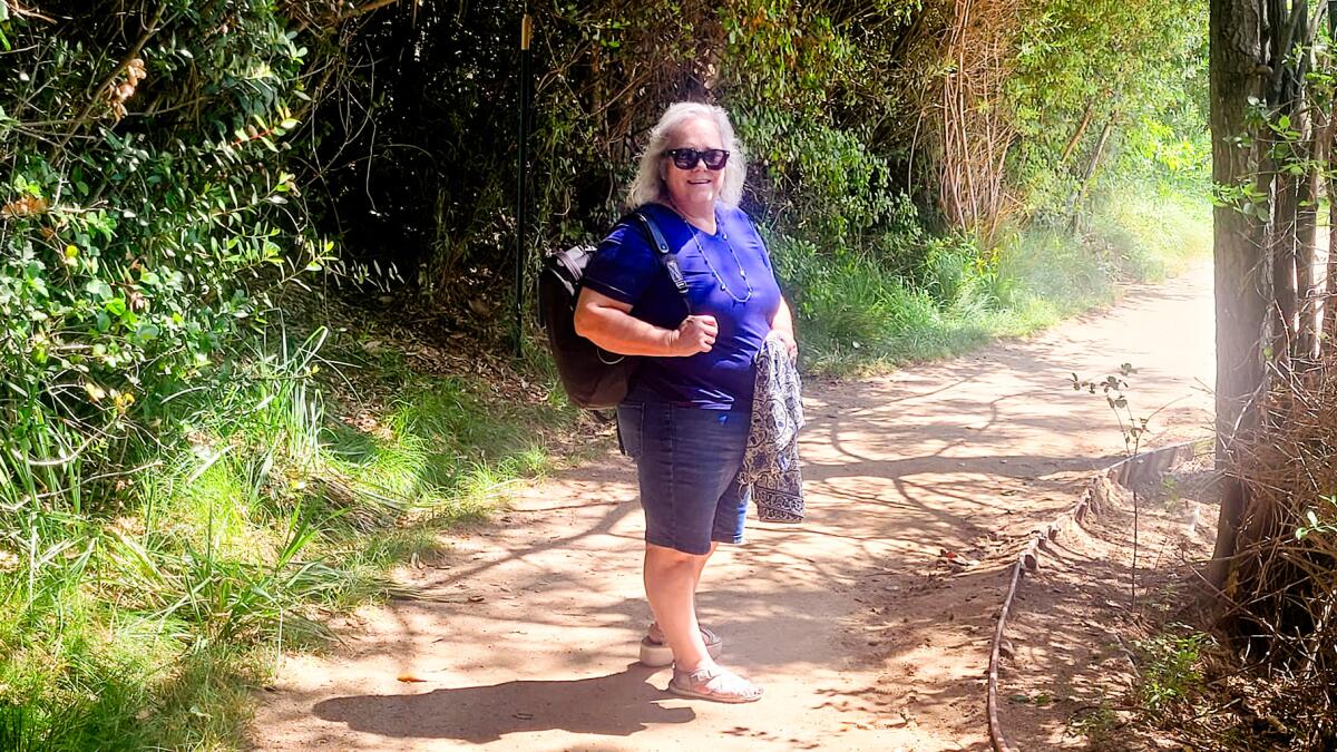 A woman stands on a dirt path among trees and shrubs