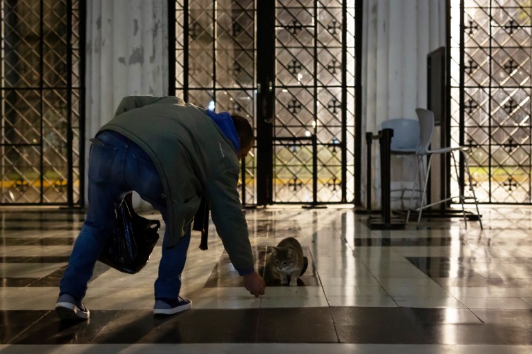 Marcelo Pisani feeds Arturito, a stray cat from outside the cemetery who visits daily at 5 a.m. to benefit from Pisani's feedings, in the entrance hall of Recoleta Cemetery in Buenos Aires, Argentina. on July 1, 2024.-Once home to a colony of more than 60 stray cats, the famed Recoleta Cemetery now houses only six cats: Lili, Princesa, Llorona, Lucio, Cabezón and Grisecito. Marcelo Pisani, the florist, visits the cemetery every day at 5am to feed the cats. However, in a country with an ever growing economic crisis and 200% inflation, he is finding the cost of looking after the animals increasingly prohibitive and has become reliant on donations from visitors. [Maria Amasanti / Al Jazeera]