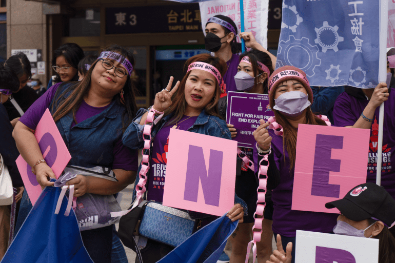 Three migrant worker women at a protest. They are wearing purple T-shorts and have bandanas around their heads. They are carrying pink placards which together spell the word 'ONE'. They are smiling.