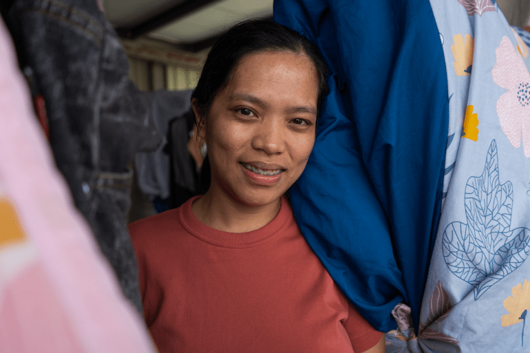 A head and shoulders portrait of Ronalyn Asis. She is wearing an orange -shirt and has long black hair. She is standing between laundry hanging from a washing line