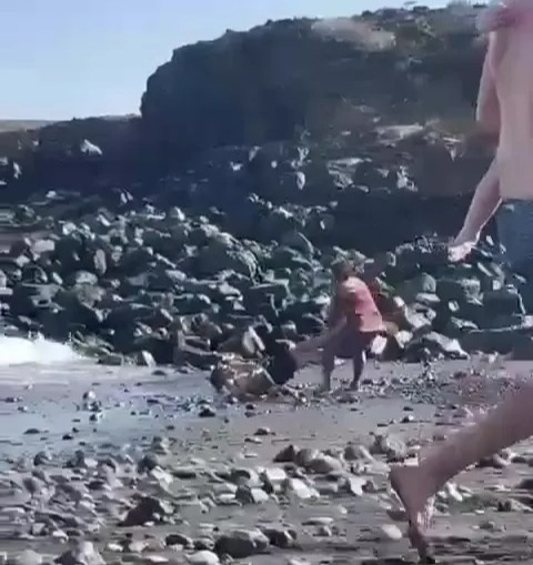 The victim rolls down the beach while the man continuously pelts him with a pole