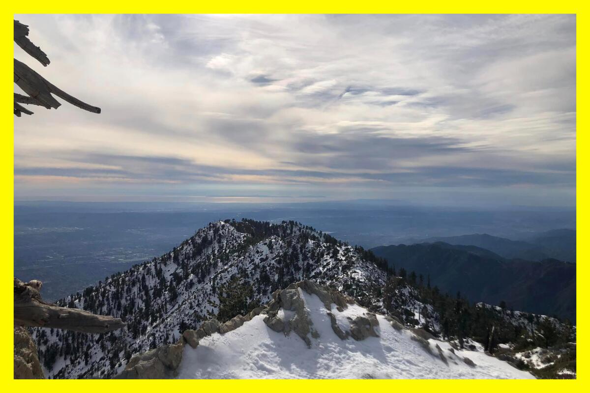 A view from Ontario Peak shows cloudy skies, snow and tree covered mountains and a vast plain in the distance.