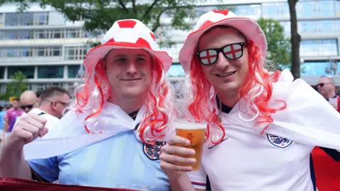 PA Two men wear England shirts and red and white hats with red and white hair attached to it