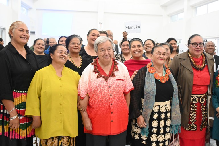 Antonio Guterres with a group of women in Tonga. He is wearing a red and white shirt with a decorative neck;ace. He is smiling. One of the women has her arm through his.