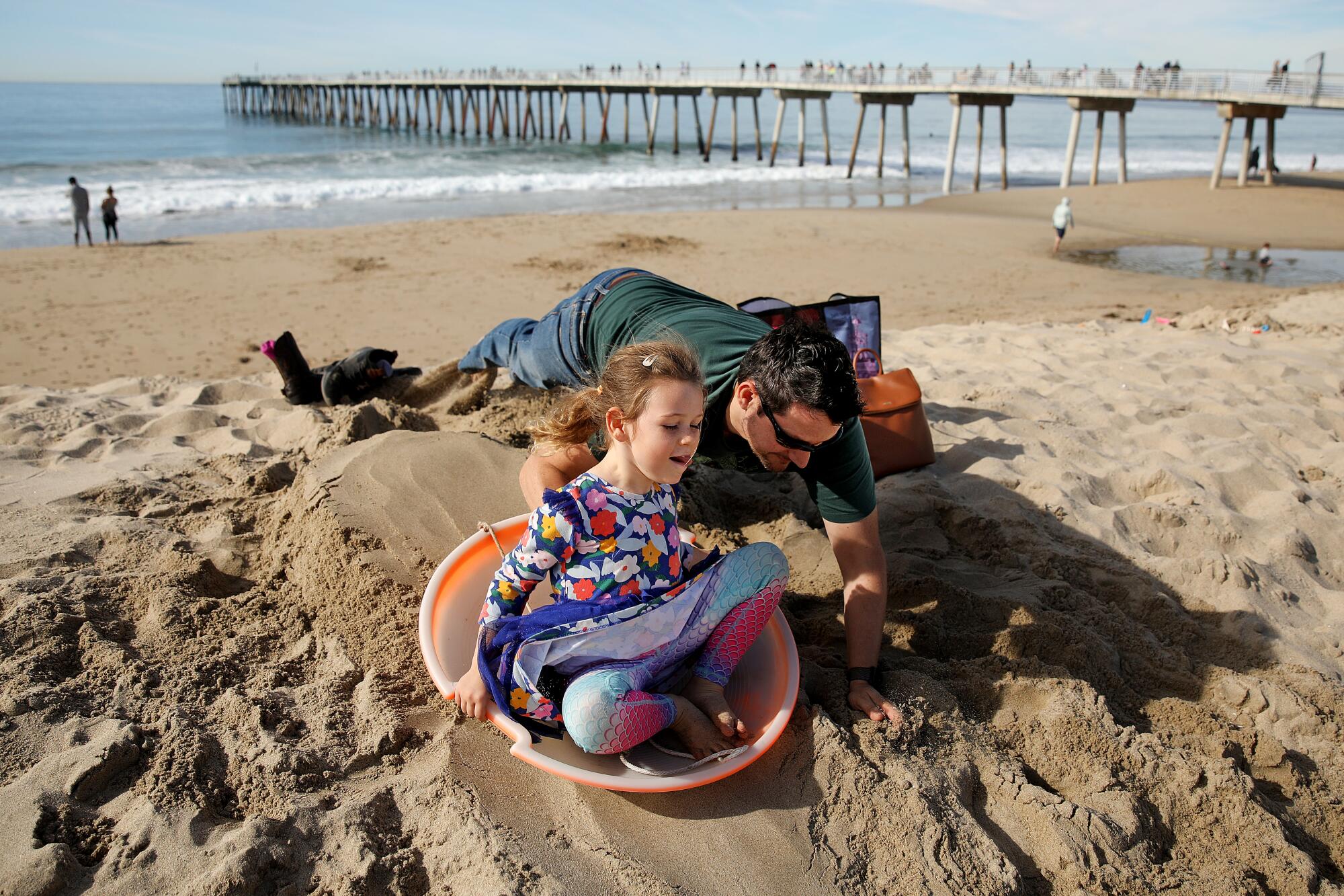 A man pushes a girl on a sled down a hill of sand.