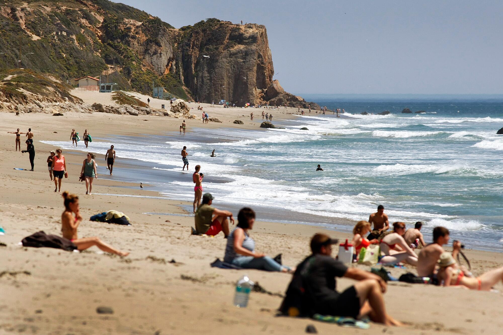 People sit on towels along the sand as white foamy waves form in the ocean. 