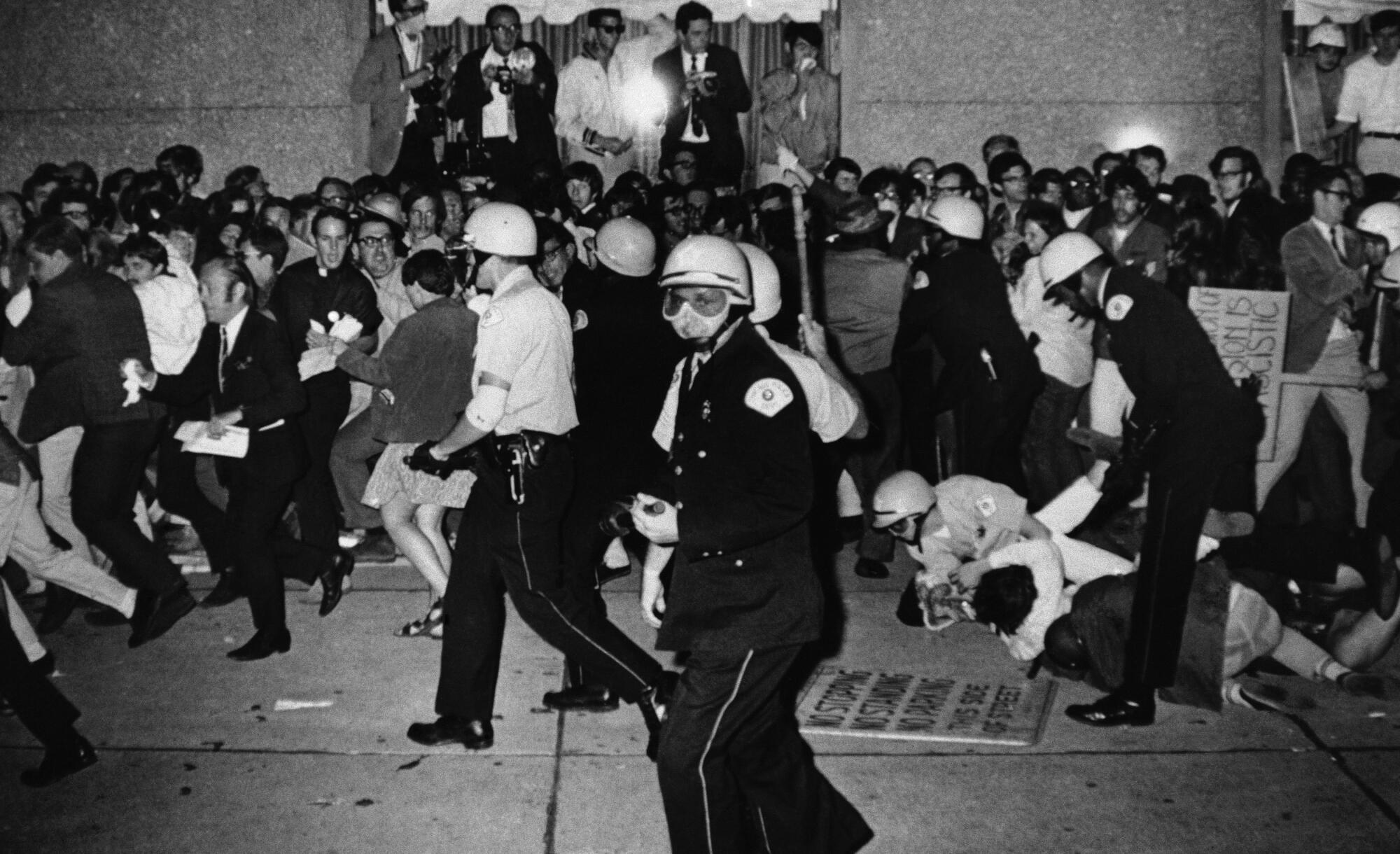 Police officers in helmets roust protesters; one officer pins a protester to the ground