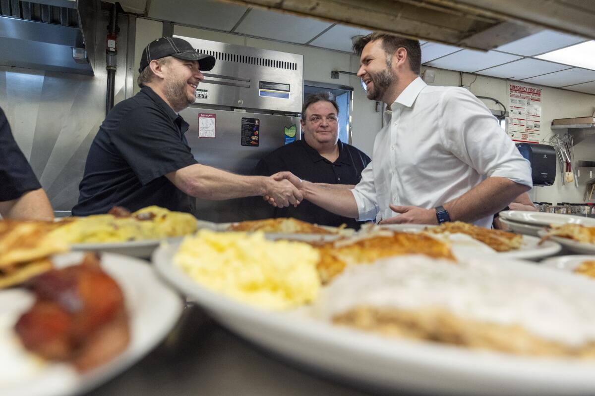 A man with dark hair and beard, in white shirtsleeves, right, shakes hands with another man near plates of food