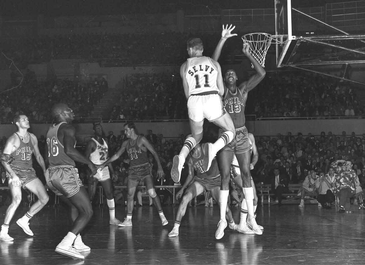 A basketball player is shown poised above the floor trying to make a basket but being blocked by a very tall player.