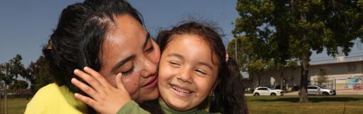 A mom kisses her her 4-year-old daughter's cheek from behind, laughing as she pushes her away.