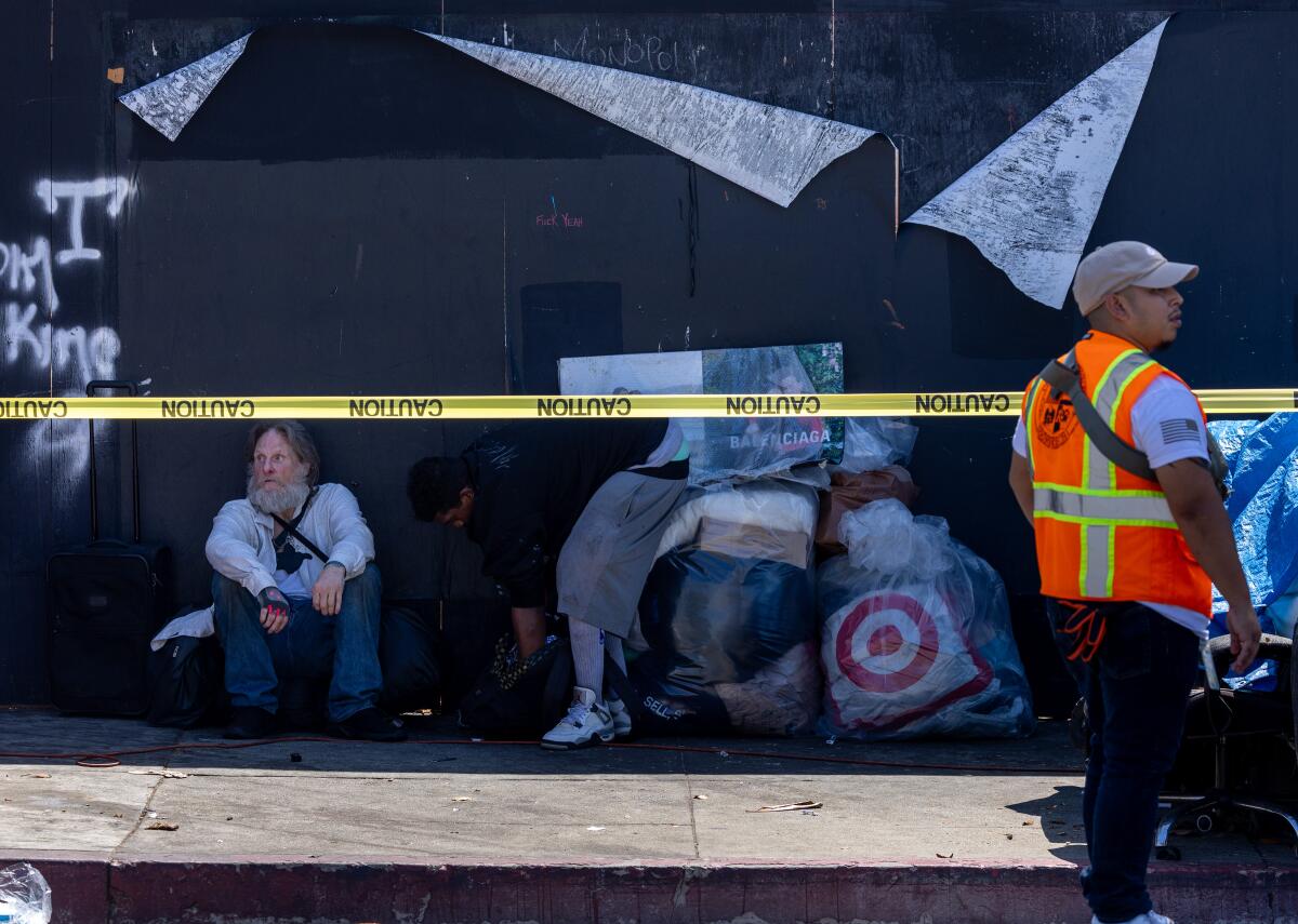 A homeless man with bags sits in the shade against a wall along the sidewalk with yellow police tape in front of it.