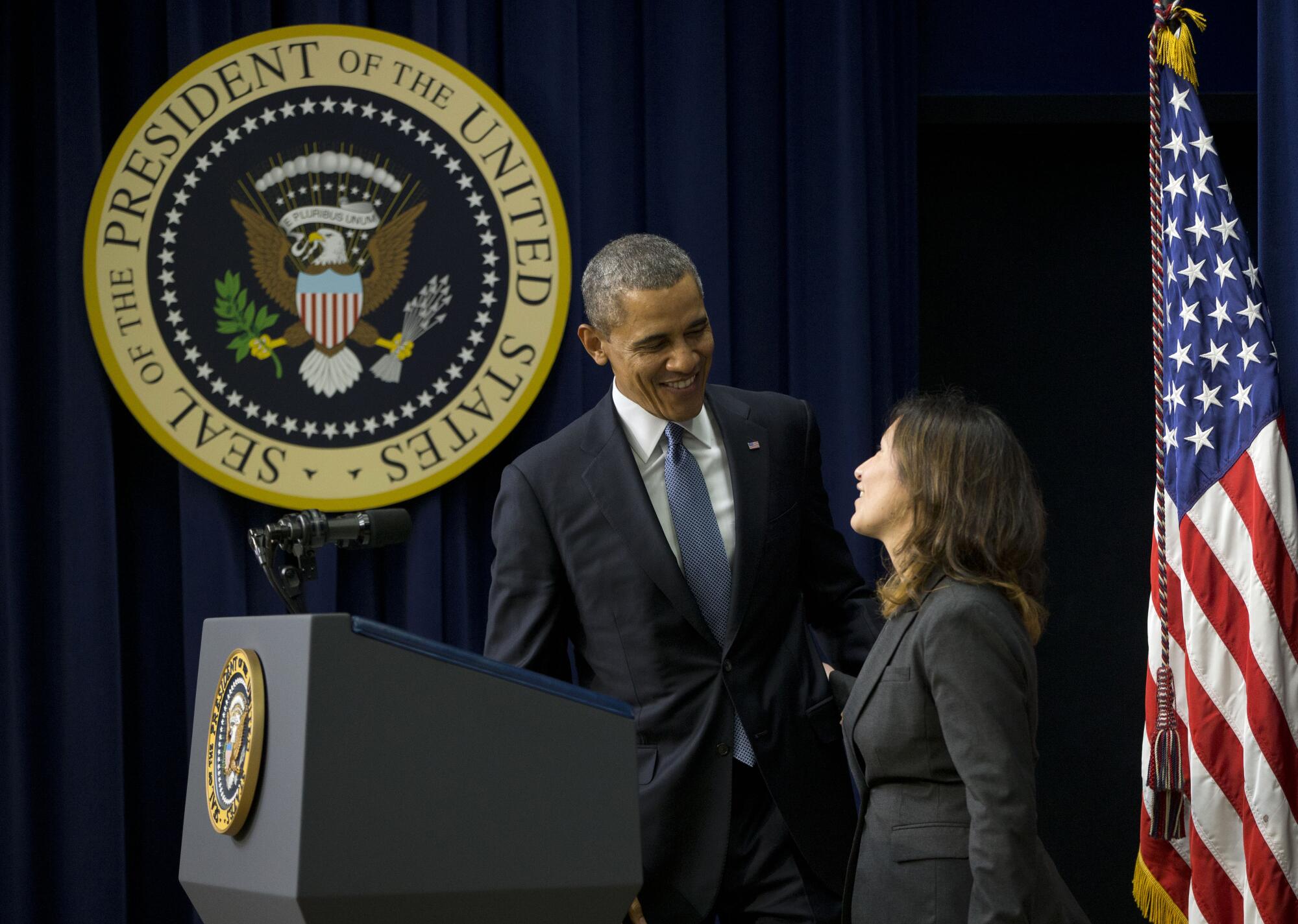 A man in dark suit and blue tie smiles at a woman, right, as they stand at a lectern, with the presidential seal behind them