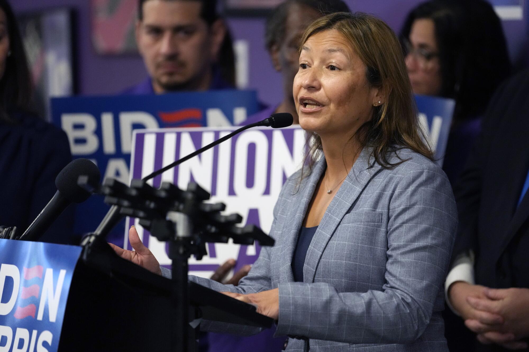 A woman with dark brown hair, wearing a gray jacket, speaks at a lectern 