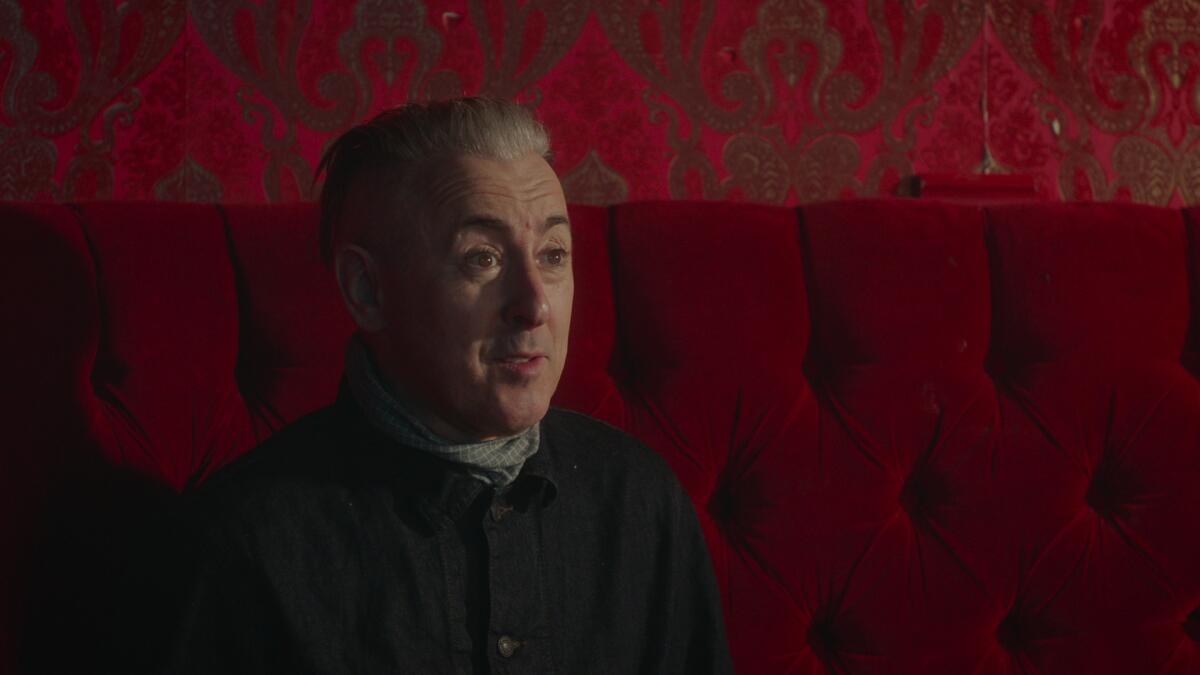 Alan Cumming in a dark shirt sitting on a red velvet couch with red wallpaper behind him.