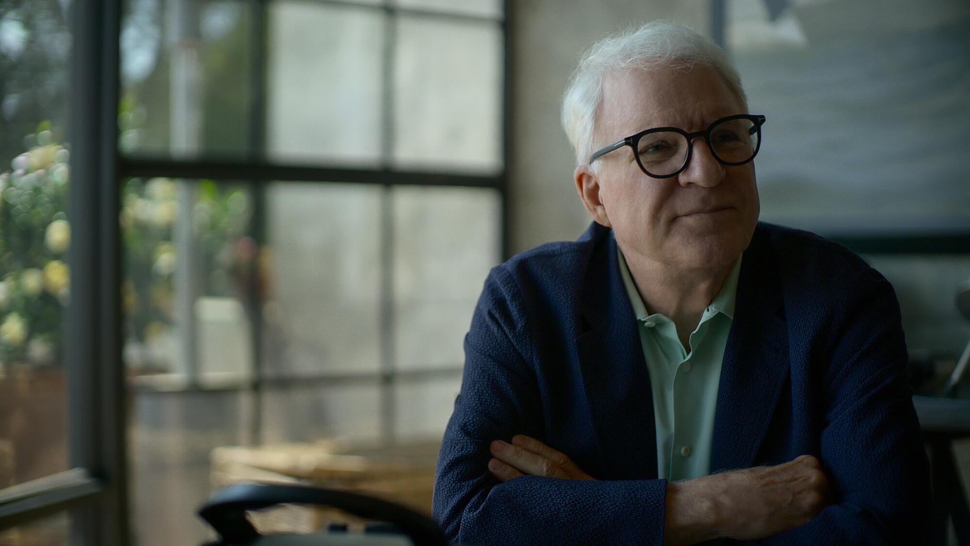 Steve Martin sits at a table and looks off at an angle.