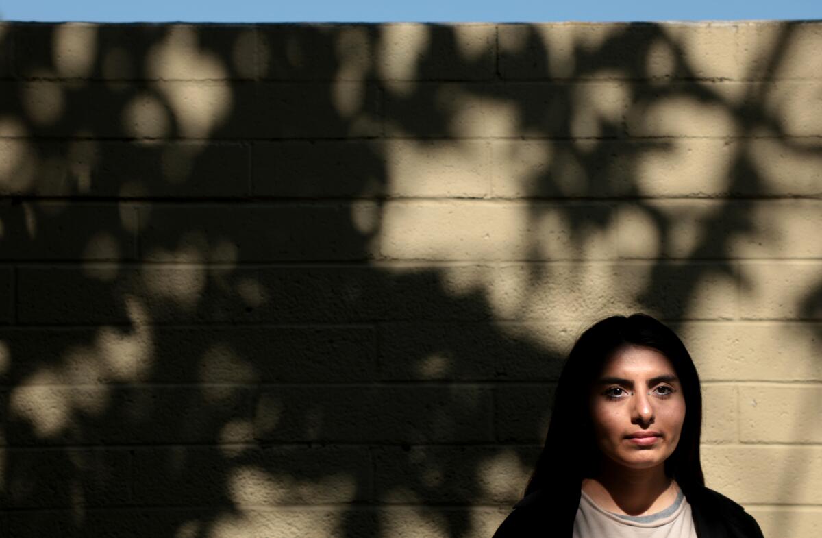 A young person with long black hair poses next to a barren brick wall.