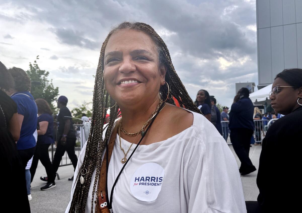 A woman with a "Harris for president" sticker on her shirt stands smiling under a cloudy sky as people pass by.