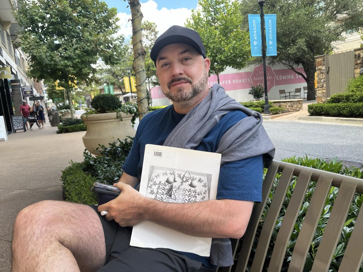 A man in a baseball cap seated on a metal bench on a sidewalk in a tree-lined retail area