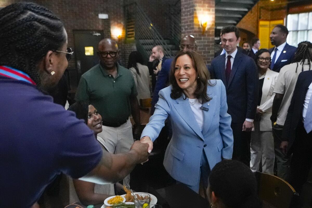 A person reaches across a table in a brick-walled eatery to shake Kamala Harris' hand as a seated women and others look on