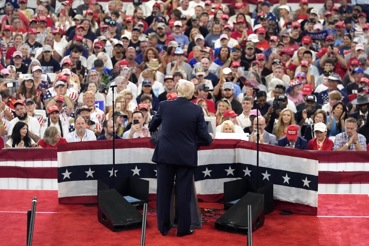 Donald Trump seen at a distance from behind, standing on a red stage decorated with stars and stripes as a crowd faces him