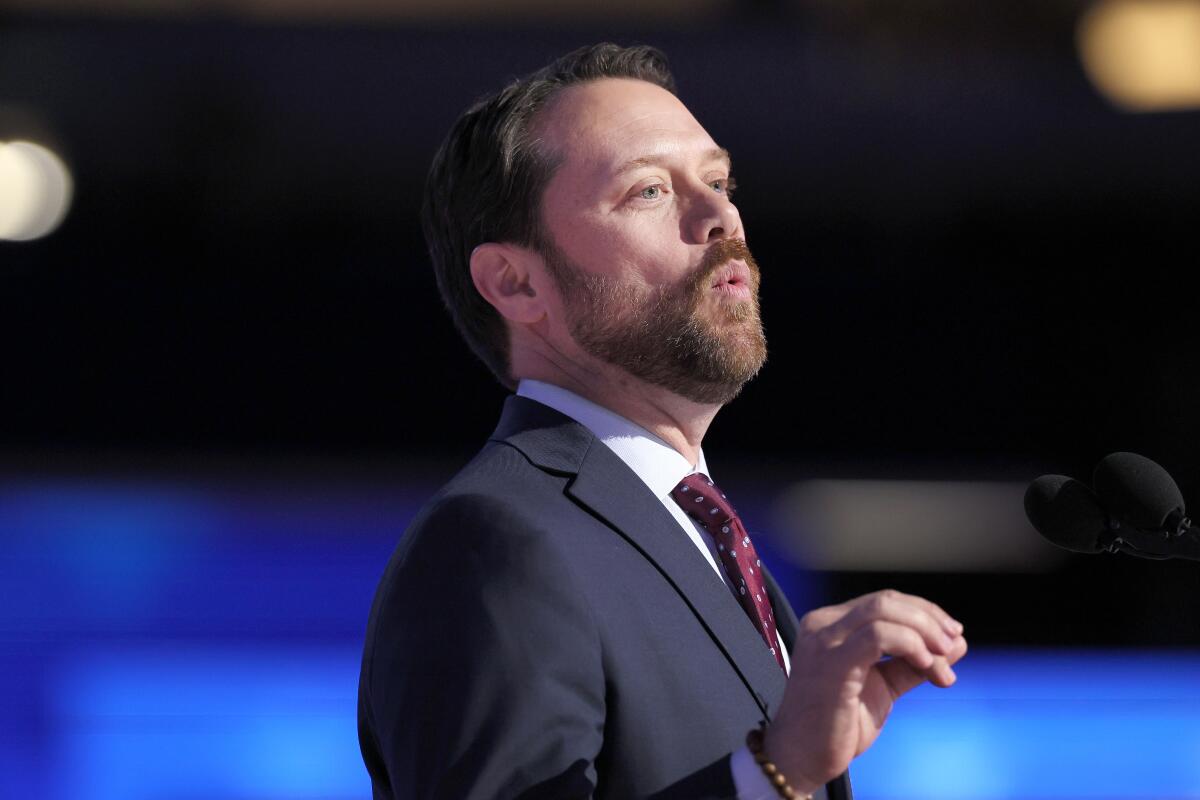 A man with dark hair and beard, in dark jacket and red tie, gestures with one hand while speaking