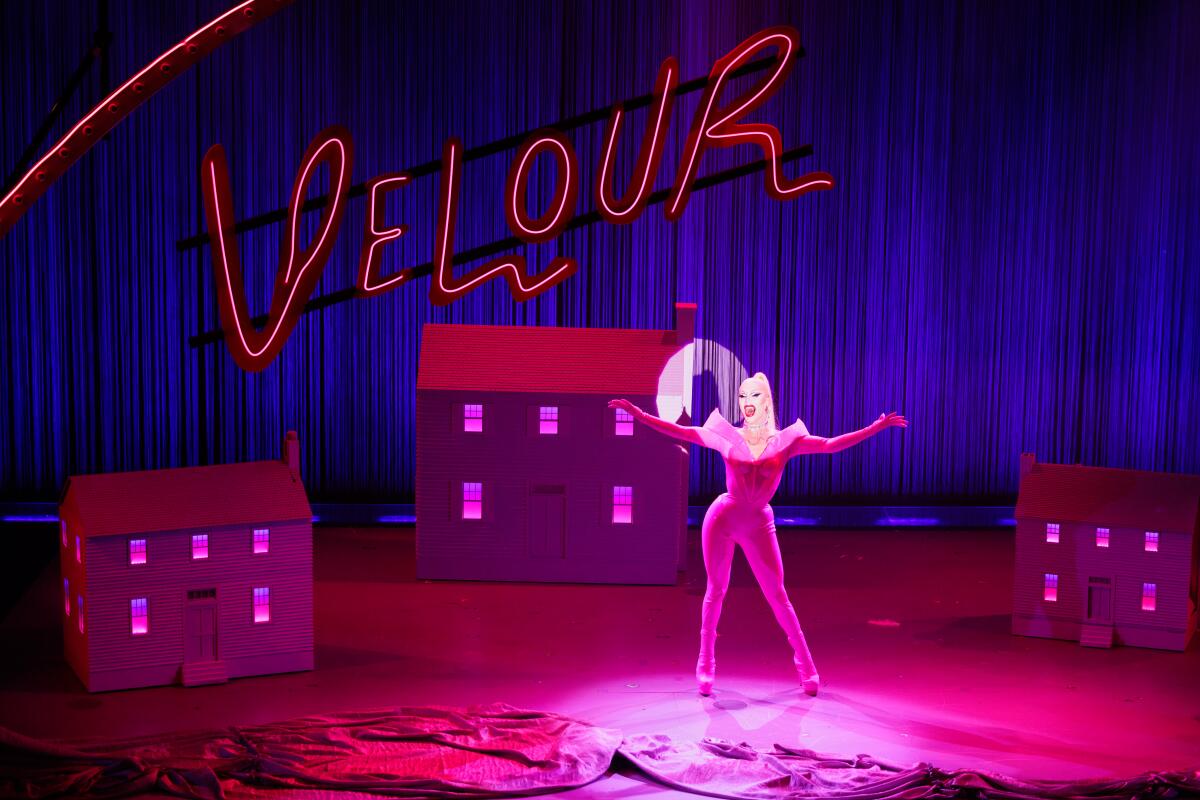 A person in a pink bodysuit surrounded by model houses and the word "Velour" in neon lights on a stage.