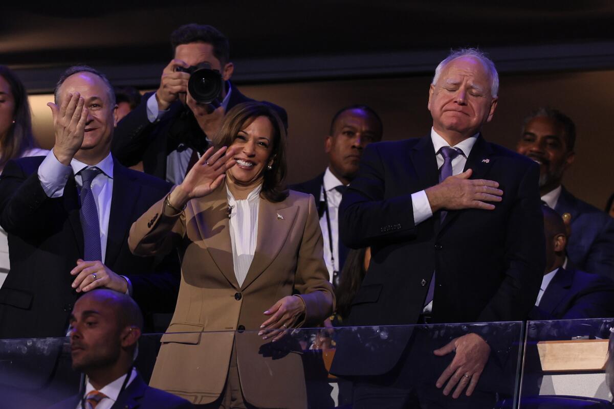 A woman in a brown jacket waves, flanked by a waving man in a dark suit, left, and another man with a hand over his chest 