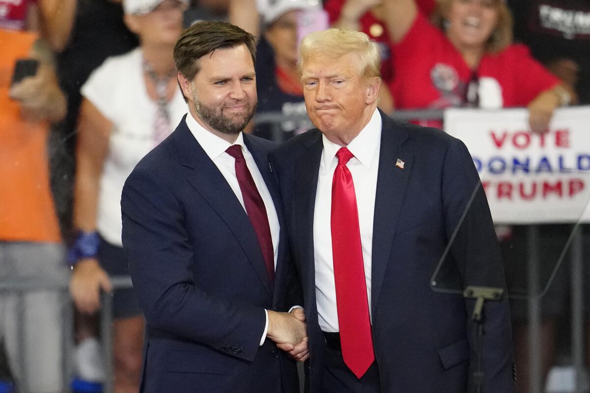 Two men, one with a beard, left, in dark suits and red ties, shake hands