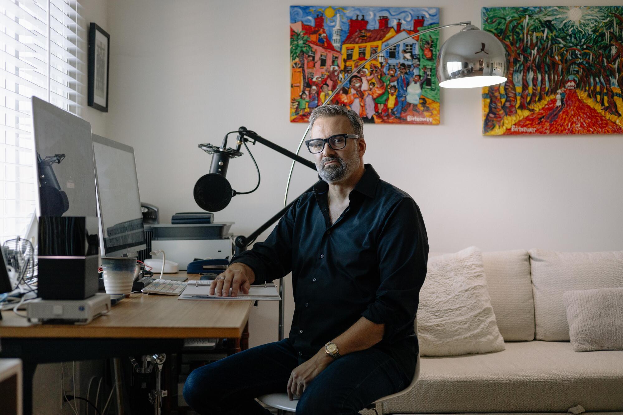 A man with salt and pepper hair wearing black rimmed glasses sits at a desk near a computer monitor.