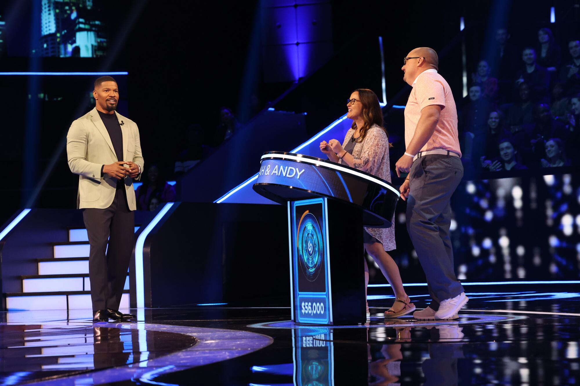 Jamie Foxx on the set of a game show looking at two contestants standing behind a podium.