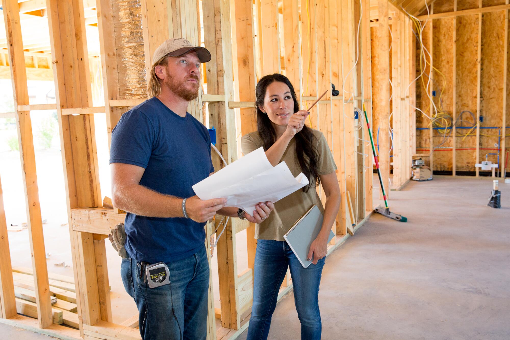 A man in a blue shirt and jeans holding a floor plan looking at the frame of a house with a woman.