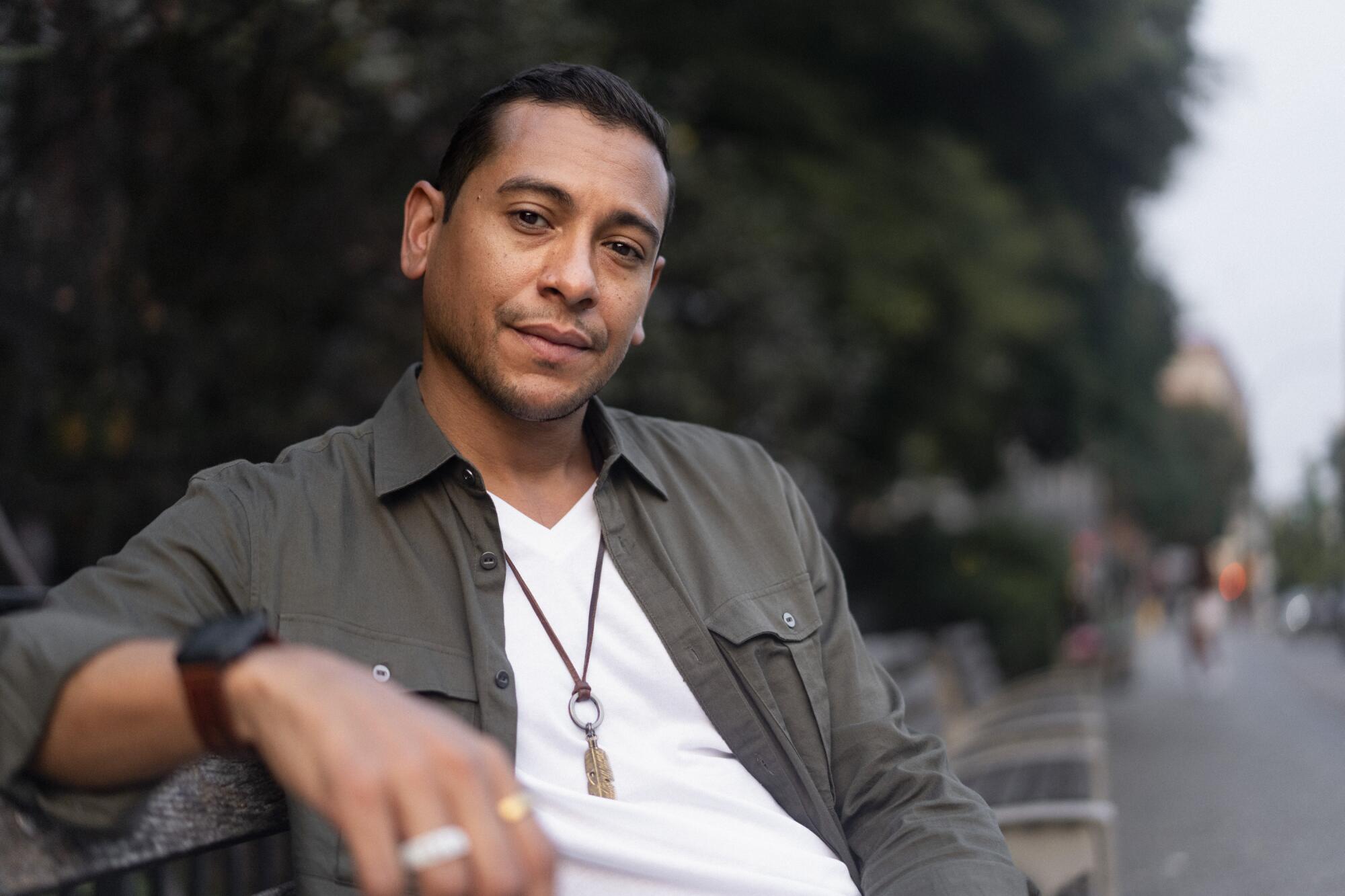 A man in a green shirt with a white T-shirt underneath sitting on a bench.