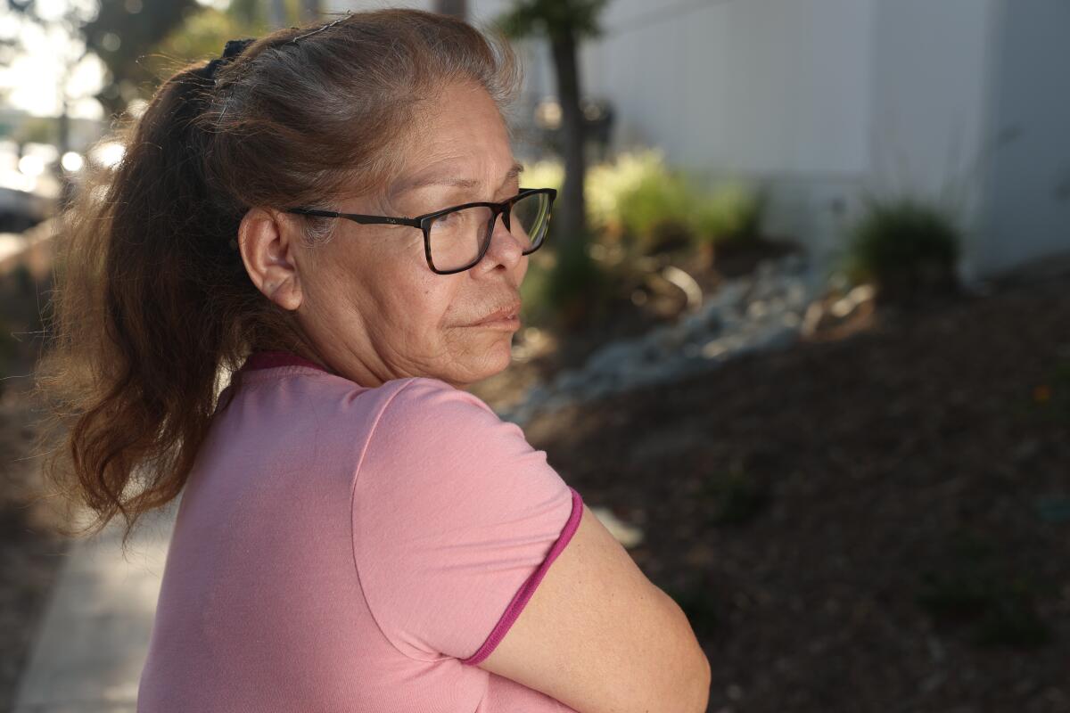 A woman in a pink T-shirt, her brown hair in a ponytail, poses on a landscaped sidewalk.  