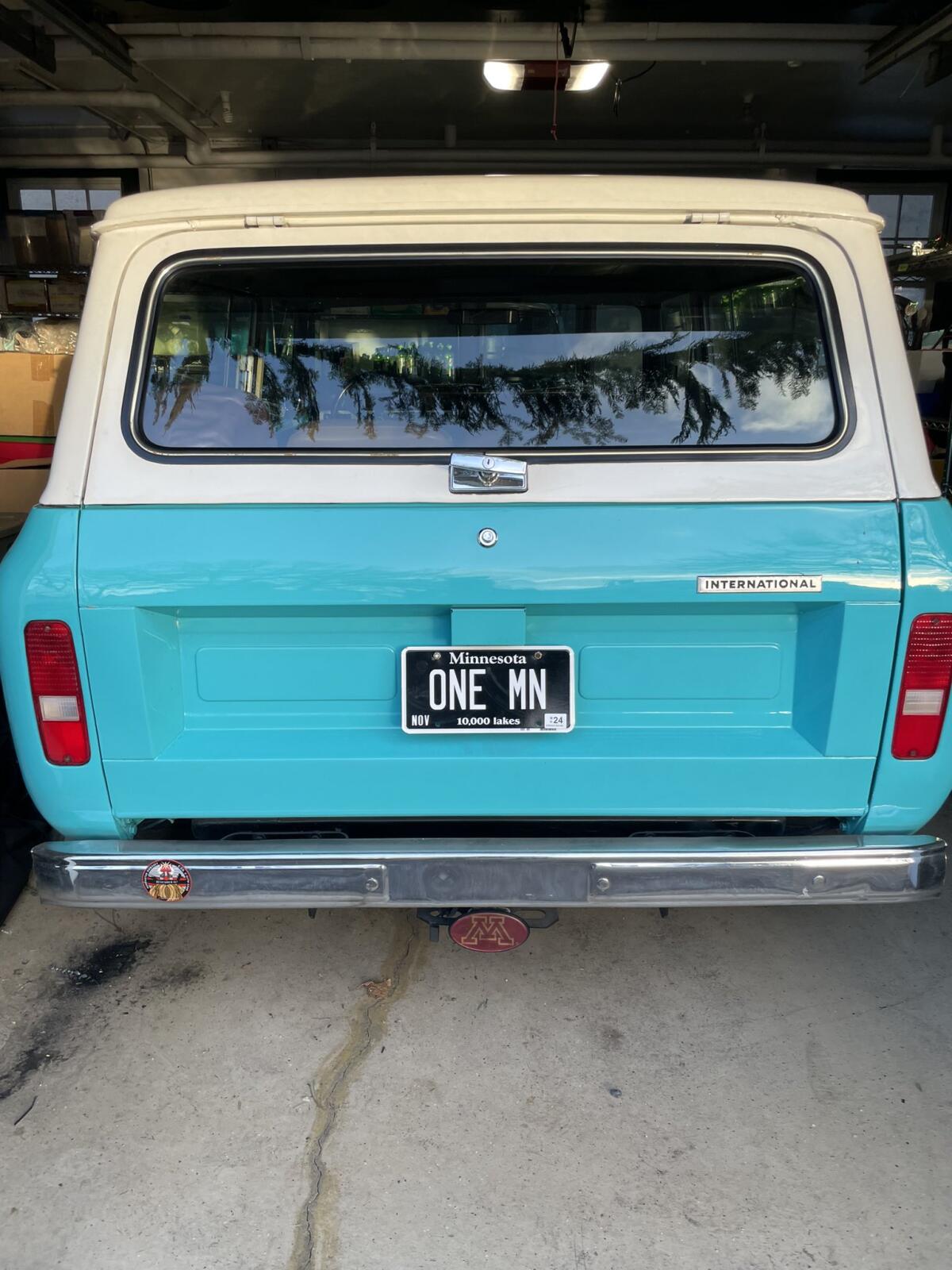 A turquoise truck with a white top and a personalized black Minnesota license plate reading "ONE MN," seen from behind.