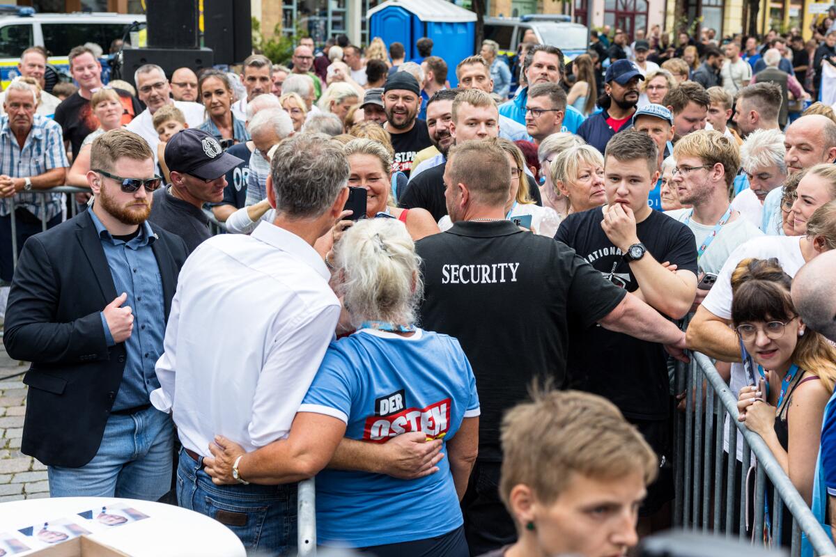 A man in a white shirt, left, and a woman in a blue shirt, with arms around each other, face a crowd 