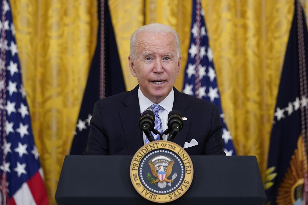 President Biden speaking from a lectern with the presidential seal against a background of flags and yellow-gold drapery