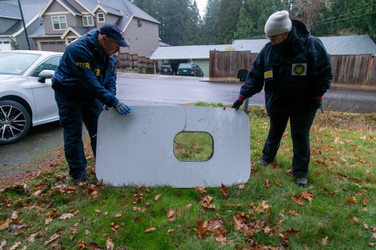 Two investigators holding up the panel that blew out of an Alaskan Airlines plane midflight. They are standing in someone's garden. The panel has a window opening