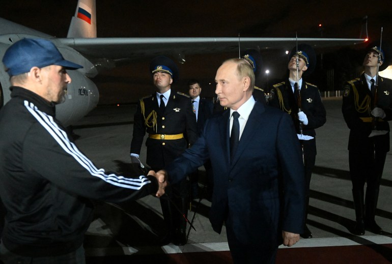 Putin greets Vadim Krasikov as he gets off the plane in Moscow. They are shaking hands. Krasikov is wearing a baseball cap and track suit. It's dark.