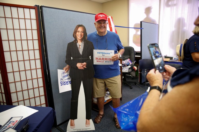 A veteran in a red cap holds up a sign that reads "Veterans for Harris" next to a cardboard cut-out of Kamala Harris.